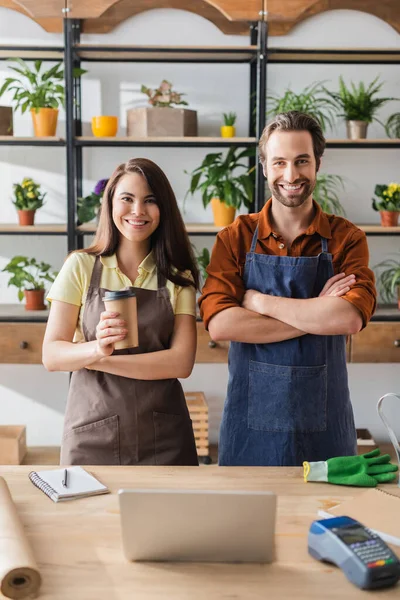 Fioristi positivi con caffè per andare a guardare la fotocamera vicino al computer portatile e terminale di pagamento nel negozio di fiori — Foto stock