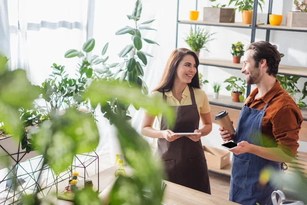 Cheerful florists with smartphone and digital tablet talking in flower shop — Stock Photo