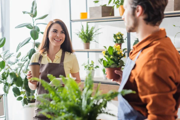Floristería sonriente sosteniendo café para llevar y tableta digital cerca de colega en floristería — Stock Photo