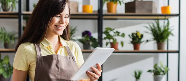 Positive florist in apron holding digital tablet in flower shop, banner — Stock Photo