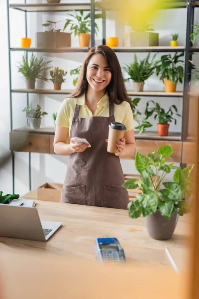 Joyeux fleuriste tenant smartphone et café pour aller dans le magasin de fleurs — Photo de stock