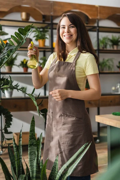 Positive florist holding sprayer near plants and looking at camera in flower shop — Stock Photo
