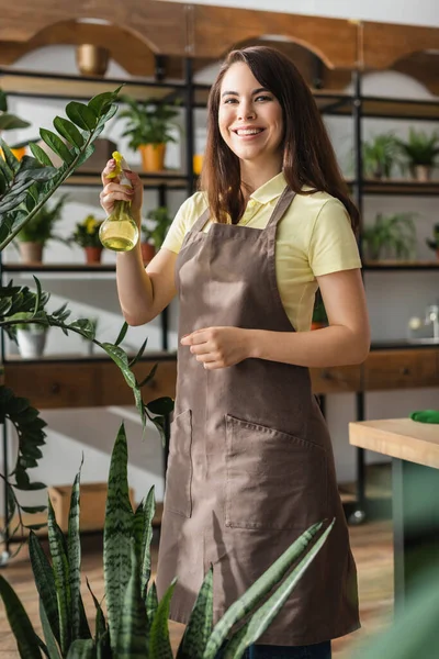 Positive Floristin hält Sprayer in der Nähe von Pflanzen und blickt in Blumenladen in die Kamera — Stockfoto