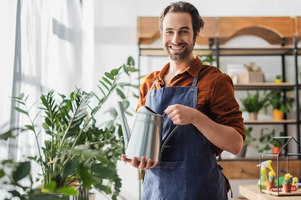 Positive florist holding watering can and looking at camera near plants in flower pot — Stock Photo