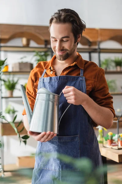 Junge Floristin in Schürze blickt in Blumenladen auf Gießkanne — Stockfoto