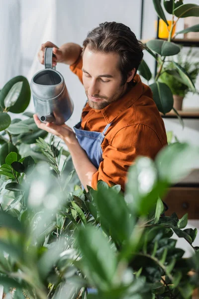 Morena florista em avental segurando rega pode perto de plantas desfocadas na loja — Fotografia de Stock