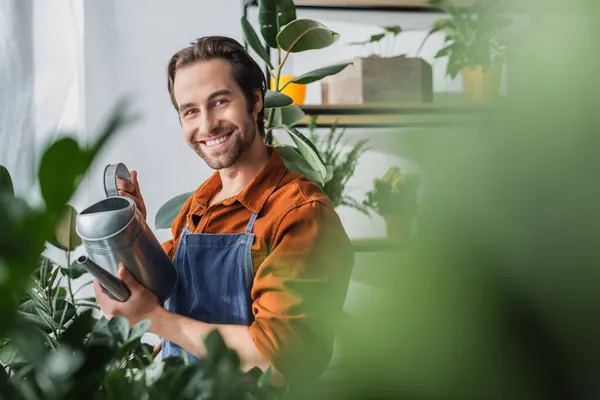 Joven florista en delantal sosteniendo regadera y mirando a la cámara cerca de plantas borrosas en la florería - foto de stock