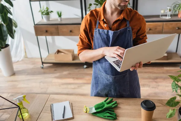 Cropped view of florist in apron using laptop near gloves, sprayer and notebook in flower shop — Stock Photo