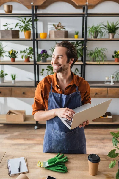 Joyeux fleuriste en tablier tenant à l'aide d'un ordinateur portable près de tasse en papier et gants dans la boutique de fleurs — Photo de stock