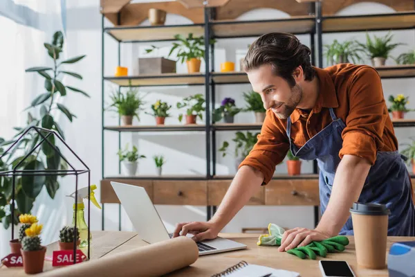 Joven vendedor en delantal usando portátil cerca de café para ir y cactus en la tienda de flores - foto de stock