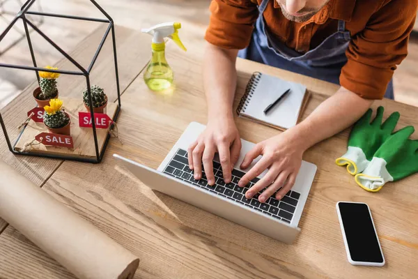 Cropped view of florist using laptop near gloves and cacti with price tags in flower shop — Stock Photo