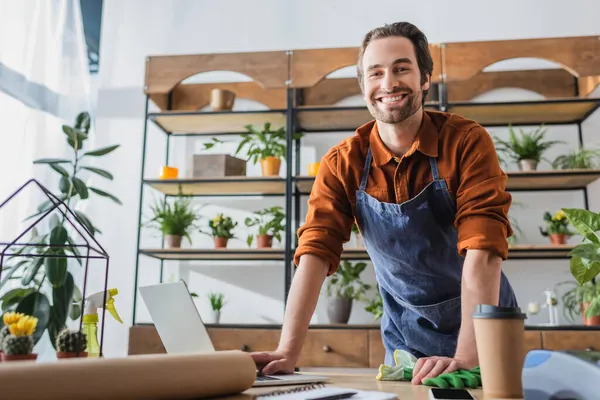 Smiling florist in apron looking at camera near gloves and laptop in flower shop — Stock Photo