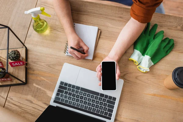 Vista dall'alto di fiorista che tiene smartphone e scrive su notebook vicino a computer portatile e cactus in negozio di fiori — Foto stock