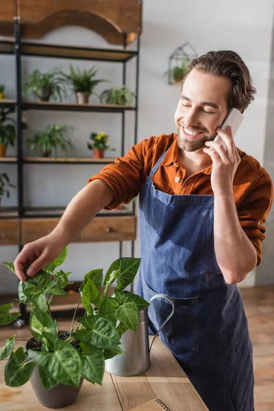 Lächelnder Florist in Schürze spricht mit Smartphone in Pflanzennähe und Gießkanne im Blumenladen — Stockfoto