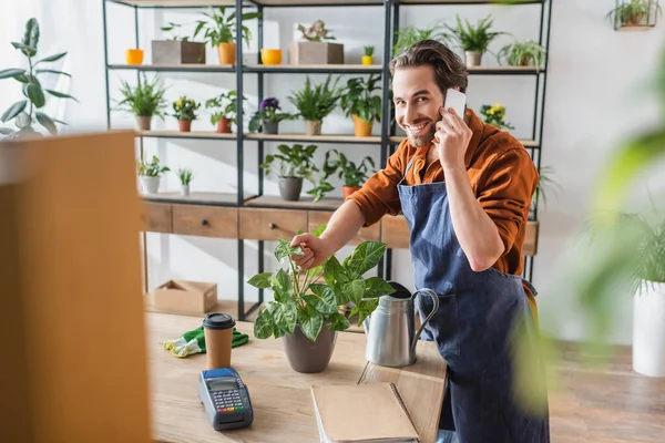 Détaillant parlant sur smartphone et touchante plante près de l'arrosoir et terminal de paiement dans le magasin de fleurs — Photo de stock