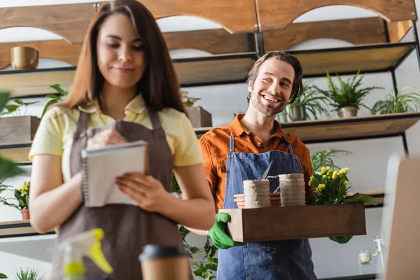 Lächelnder Blumenhändler hält Box mit Blumentöpfen neben Kollegin mit Notizbuch im Blumenladen — Stockfoto