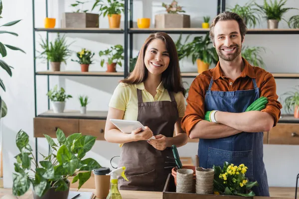 Positive florists in aprons with digital tablet looking at camera near plants and coffee in flower shop — Stock Photo