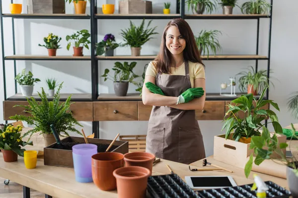 Florista sorridente em luvas olhando para a câmera perto de tablet digital e ferramentas de jardinagem na loja de flores — Fotografia de Stock