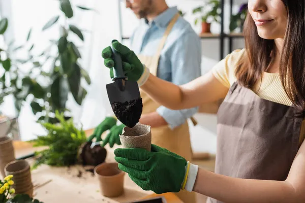 Cropped view of florist in apron pouring ground in flowerpot near blurred colleague in flower shop — Stock Photo