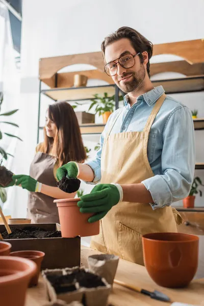 Floristería joven en anteojos y delantal vertiendo tierra cerca de macetas en floristería - foto de stock