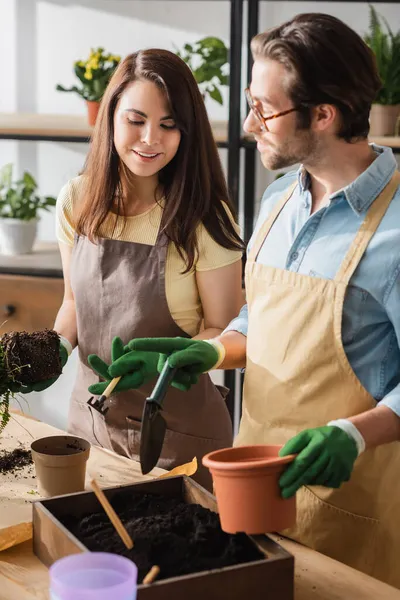 Lächelnder Blumenhändler mit Harke blickt auf Boden neben Kollege zeigt mit Finger in Blumenladen — Stockfoto