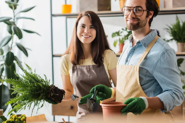 Young florist in eyeglasses holding shovel and flowerpot near cheerful colleague in flower shop — Stock Photo