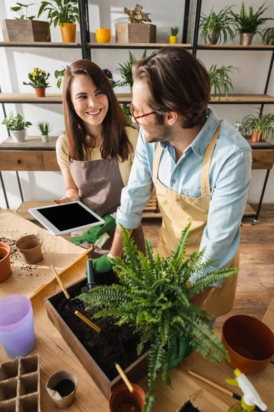 Lächelnder Blumenhändler hält digitales Tablet in der Nähe von Kollegen und Gartengeräten im Blumenladen — Stockfoto