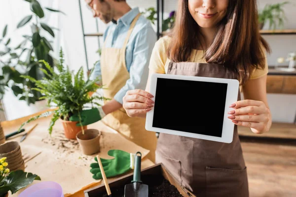 Fiorista in grembiule con tablet digitale con schermo bianco vicino collega sfocato e piante in negozio di fiori — Foto stock