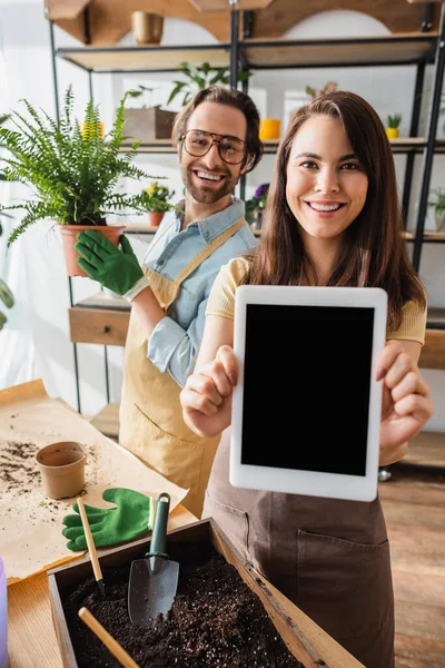 Fleuristes souriants avec plante et tablette numérique regardant la caméra près des outils de jardinage dans le magasin de fleurs — Photo de stock