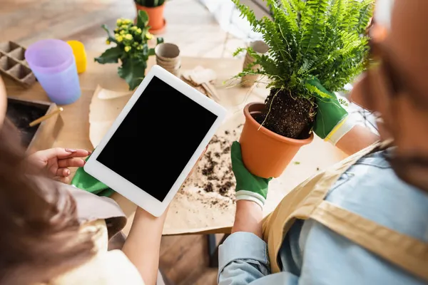 Fiorista offuscata che tiene la targa digitale vicino a collega con vaso da fiori e impianto in negozio — Foto stock