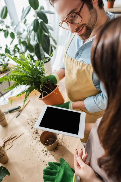 Joven florista sosteniendo tableta digital con pantalla en blanco cerca de colega sonriente con planta en floristería - foto de stock