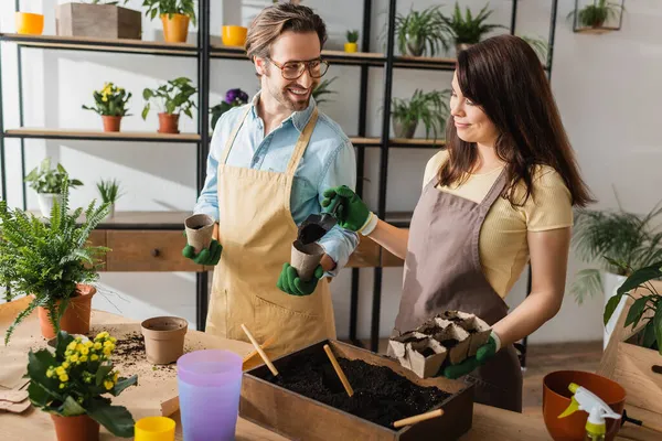 Smiling florist pouring ground near colleague with flowerpots in flower shop — Stock Photo