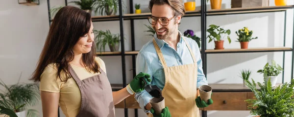 Florist pouring ground near positive colleague with flowerpots in shop, banner — Stock Photo