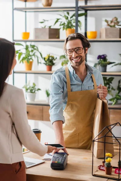 Floristería sonriente sosteniendo bolsa de compras cerca de la terminal de pago y cliente borroso con teléfono inteligente en la tienda de flores - foto de stock