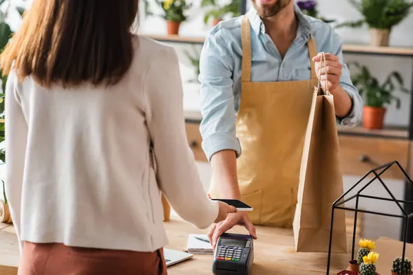 Vista ritagliata del cliente che paga con smartphone vicino fiorista con shopping bag nel negozio di fiori — Foto stock