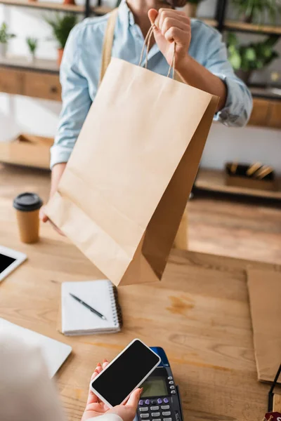 Vista recortada de floristería sosteniendo bolsa de compras cerca del cliente pagando con teléfono inteligente en tienda de flores - foto de stock