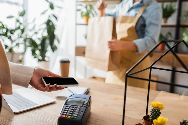 Vista recortada del cliente pagando con smartphone cerca de floristería borrosa con bolsa de compras en floristería - foto de stock