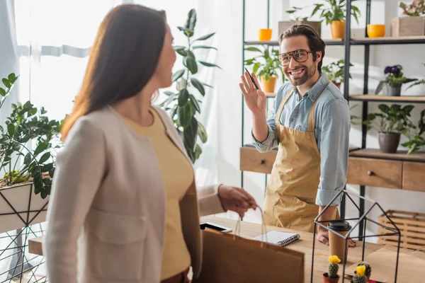 Smiling florist waving hand near blurred customer with shopping bag in flower shop — Stock Photo