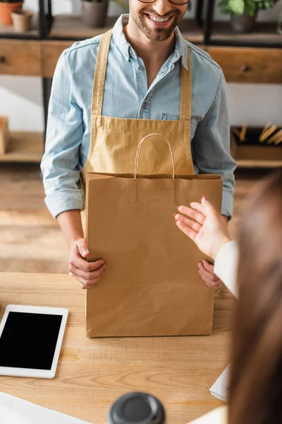 Усміхнений продавець в фартусі тримає сумку біля розмитого клієнта і цифровий планшет у квітковому магазині — стокове фото