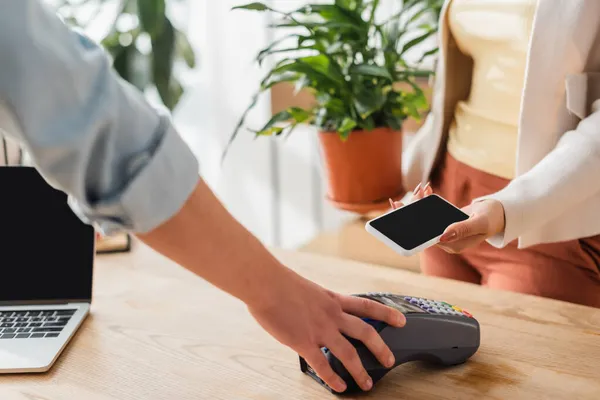 Vista cortada de florista segurando terminal de pagamento perto do cliente com smartphone na loja de flores — Fotografia de Stock