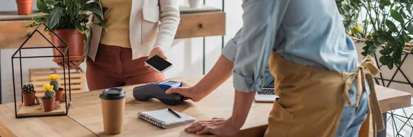 Cropped view of customer holding plant and paying with cellphone near florist and coffee in shop, banner — Stock Photo