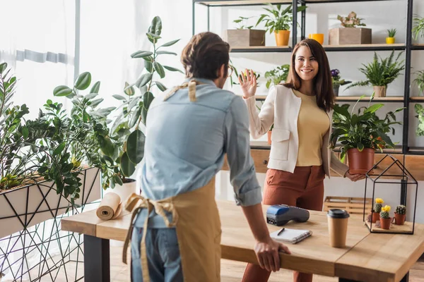 Cliente sorridente con pianta che ondeggia mano vicino fiorista e terminale di pagamento nel negozio di fiori — Foto stock