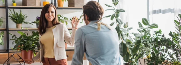 Cheerful woman holding plant and waving hand at florist in flower shop, banner — Stock Photo