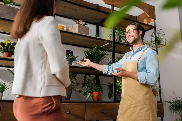 Low angle view of smiling florist pointing at plants near blurred customer in flower shop — Stock Photo