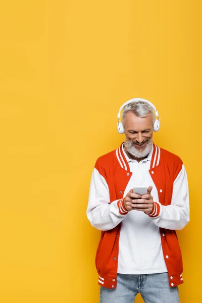 Positive middle aged man in wireless headphones holding smartphone and listening music isolated on yellow — Stock Photo