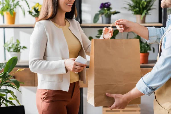 Vista recortada de un cliente sonriente sosteniendo un teléfono inteligente cerca de la floristería con bolsa de compras en la tienda de flores - foto de stock