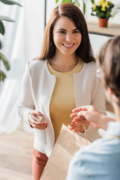 Cheerful customer holding smartphone near blurred florist with shopping bag in flower shop — Stock Photo