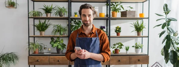 Fleuriste tenant smartphone et regardant la caméra dans le magasin de fleurs, bannière — Photo de stock