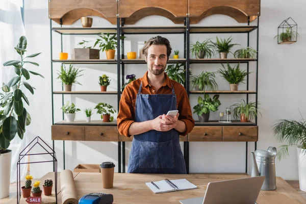 Florist holding smartphone near laptop and coffee in flower shop — Stock Photo