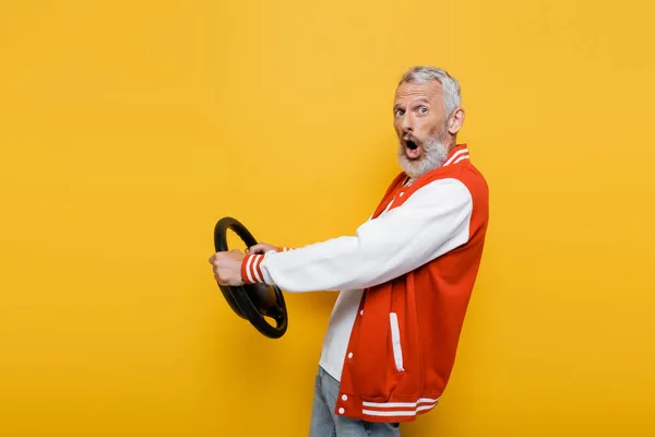 Shocked middle aged man in bomber jacket holding steering wheel while imitating driving on yellow — Stock Photo